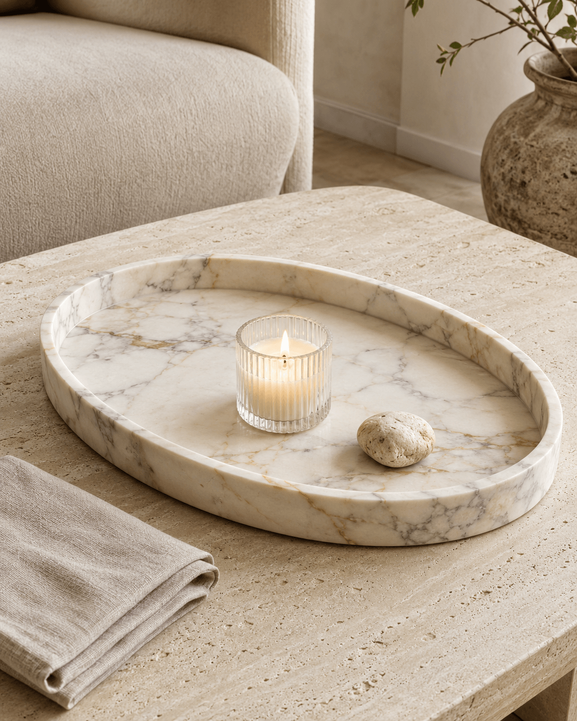 White marble tray with a glass candle and folded neutral linen on a pale stone table